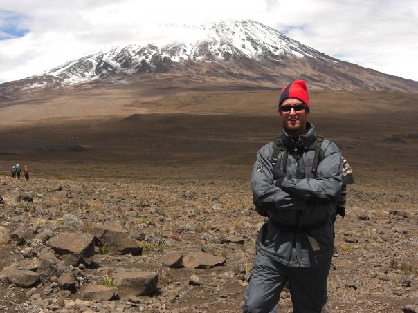 MP Turnbull dressed in a jacket, hat, and backpack, smiles with his arms crossed. Mount Kilimanjaro is in the background.